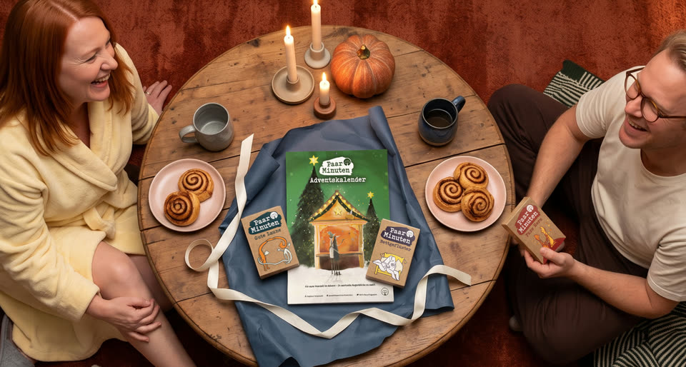 Ein Paar sitzt lachend an einem runden Holztisch mit Kerzen, Kürbis und Zimtschnecken. In der Mitte liegt der PaarMinuten Adventskalender 2026, daneben die drei Klassiker-Kartensets Fürs Herz, Gute Laune und Bettgeflüster.
