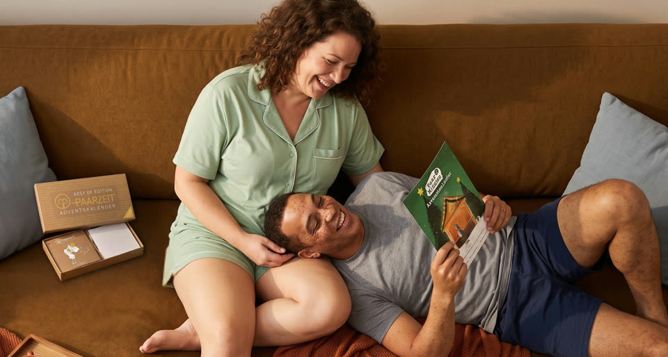Ein Paar liegt lachend auf dem Sofa: Sie sitzt im mintgruenen Pyjama, er hat den Kopf in ihrem Schoss und liest eine Karte aus dem PaarMinuten Adventskalender. Daneben steht die geoeffnete Best of Edition des Paarzeit Adventskalenders.