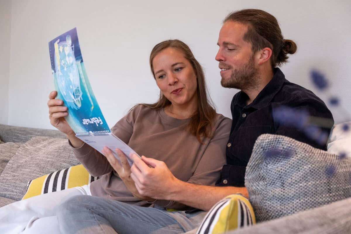 Paar sitzt auf dem Sofa und betrachtet gemeinsam ein illustriertes Monatsblatt des Paarzeit-Wandkalenders