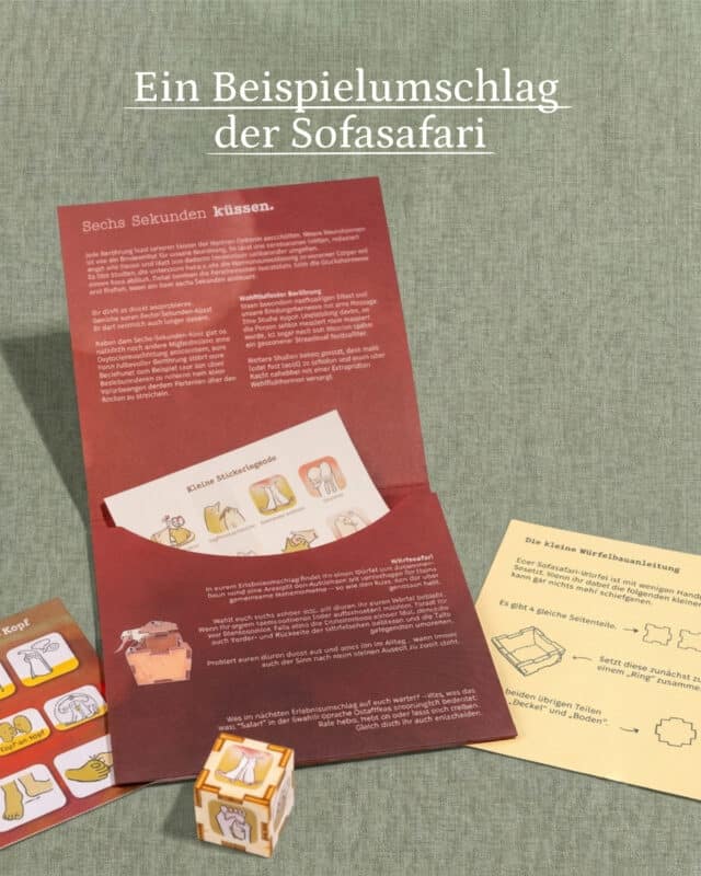 Aufgeklappter roter Erlebnisumschlag der Sofasafari mit dem Text „Sechs Sekunden küssen“, daneben Stickerbogen, Würfelbauanleitung und ein zusammengesteckter Holzwürfel auf graugrünem Leinen
