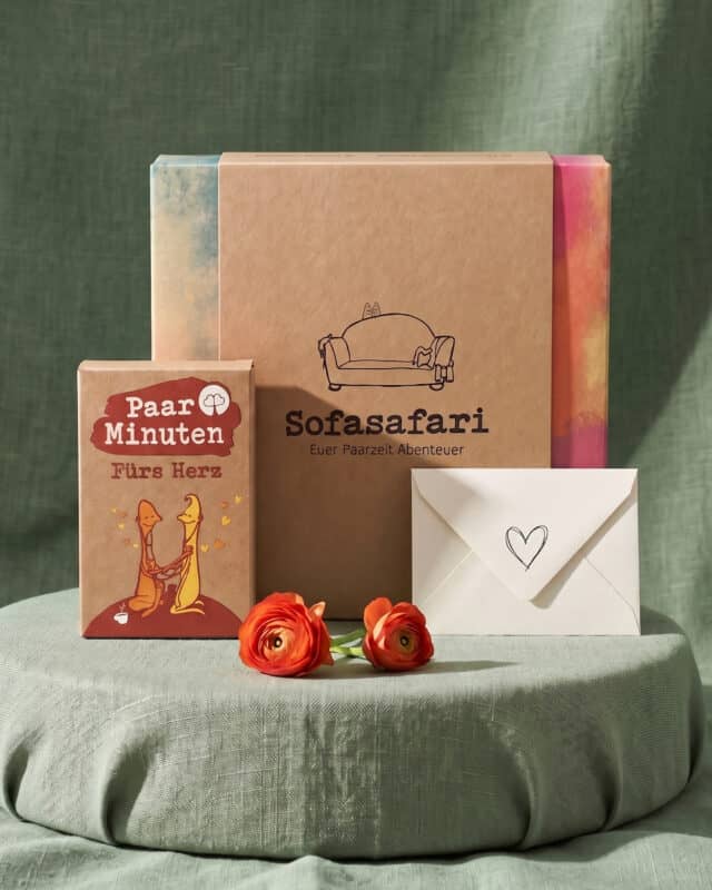Die Erlebnisbox Sofasafari mit dem Kartenset PaarMinuten Fürs Herz und einem Briefumschlag mit Herz, davor orange Ranunkeln auf grünem Leinen
