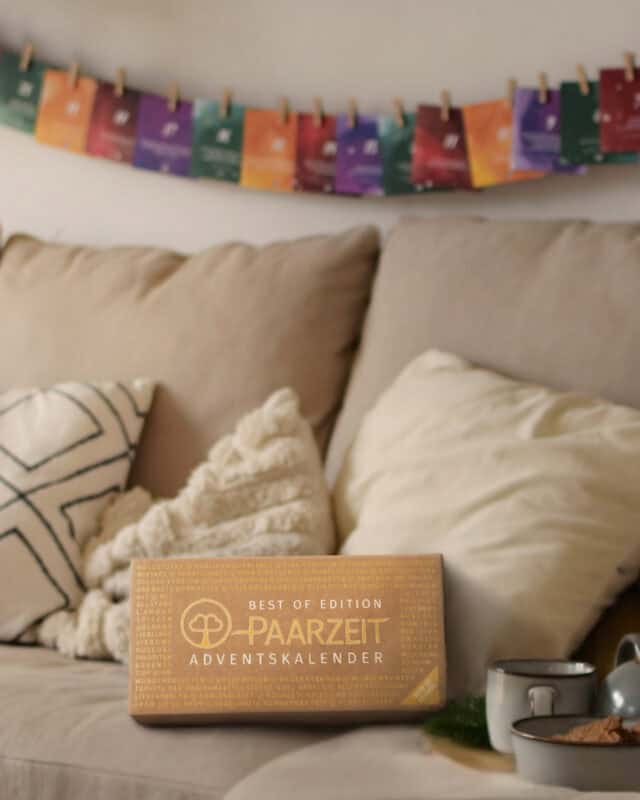 Die Kraftpapier-Schachtel des Paarzeit Adventskalenders BEST OF EDITION steht auf einem hellen Sofa mit Kissen und einer weißen Kuscheldecke, daneben zwei Tassen und Gebäck auf einem Tablett. Über dem Sofa hängen bunte Karten in Grün, Orange, Beere und Violett mit Holzklammern an einer Schnur. Auf der Schachtel in goldener Prägung: „BEST OF EDITION“, das Paarzeit-Baumlogo und „PAARZEIT ADVENTSKALENDER“, ringsum eine goldene Wortcollage.