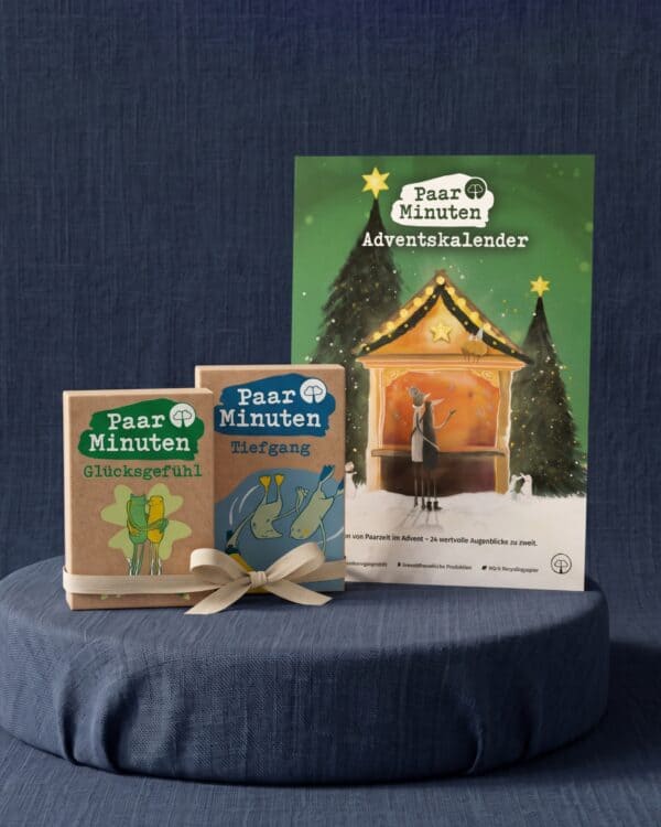 Der grüne PaarMinuten Adventskalender 2026 mit illustrierter Weihnachtsmarktbude zwischen verschneiten Tannen steht auf einem runden, blau bezogenen Podest vor dunkelblauem Stoff; davor lehnen die beiden Kartensets PaarMinuten Glücksgefühl und PaarMinuten Tiefgang in Kraftpapier-Schachteln aneinander, zusammengehalten von einer beigen Stoffschleife.