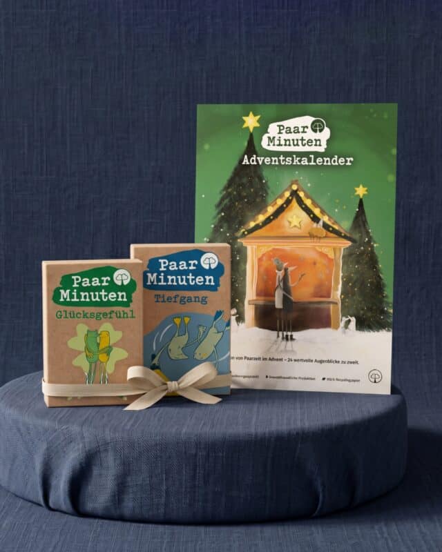 Der grüne PaarMinuten Adventskalender 2026 mit illustrierter Weihnachtsmarktbude zwischen verschneiten Tannen steht auf einem runden, blau bezogenen Podest vor dunkelblauem Stoff; davor lehnen die beiden Kartensets PaarMinuten Glücksgefühl und PaarMinuten Tiefgang in Kraftpapier-Schachteln aneinander, zusammengehalten von einer beigen Stoffschleife.