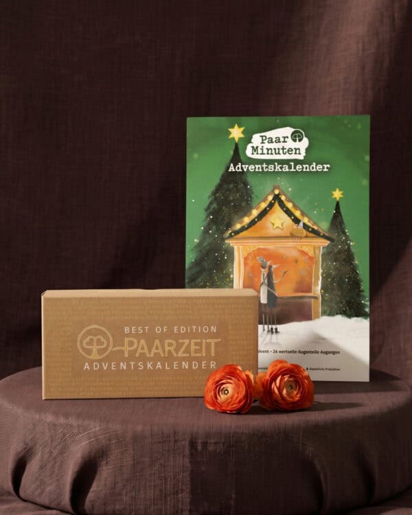 Vor dunkelbraunem Stoff steht auf einem runden, braun bezogenen Podest der grüne PaarMinuten Adventskalender mit illustrierter Weihnachtsmarktbude zwischen verschneiten Tannen und einem gelben Stern an der Spitze; davor liegt die Kraftpapier-Schachtel des Paarzeit Adventskalenders BEST OF EDITION mit goldener Prägung, daneben zwei orangefarbene Ranunkelblüten.
