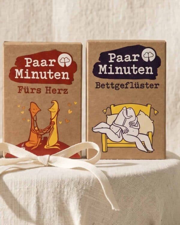 Die Kartensets PaarMinuten Fürs Herz und PaarMinuten Bettgeflüster stehen nebeneinander auf einem hellen Leinenkissen, davor eine gebundene Geschenkschleife