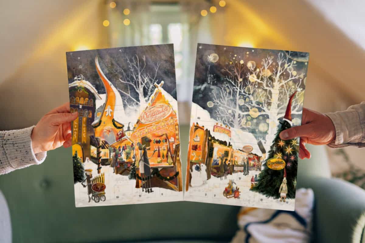 Zwei Hände halten die beiden Hälften des PaarMinuten Adventskalenders 2026 nebeneinander hoch. Beide Hälften zeigen einen gemalten Weihnachtsmarkt bei Nacht: links eine hell erleuchtete Marktbude mit der Aufschrift „Winterzauber“, daneben ein Lebkuchenhaus und Menschen zwischen den Ständen; rechts verschneite Bäume mit Lichterglanz, ein geschmückter Tannenbaum, ein Wichtel mit roter Zipfelmütze und ein Schneemann. Die Türchen sind als nummerierte, gestrichelte Felder angedeutet.