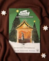 Der grüne PaarMinuten Adventskalender 2026 liegt auf braunem Strickstoff zwischen Zimtsternen; die Illustration zeigt ein Paar vor einer beleuchteten Weihnachtsmarktbude zwischen verschneiten Tannen