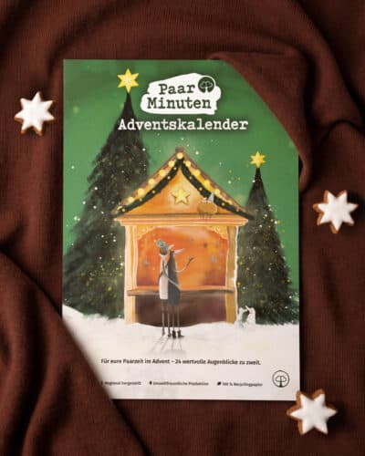 Der grüne PaarMinuten Adventskalender 2026 liegt auf braunem Strickstoff zwischen Zimtsternen; die Illustration zeigt ein Paar vor einer beleuchteten Weihnachtsmarktbude zwischen verschneiten Tannen, darunter „Für eure Paarzeit im Advent – 24 wertvolle Augenblicke zu zweit“.
