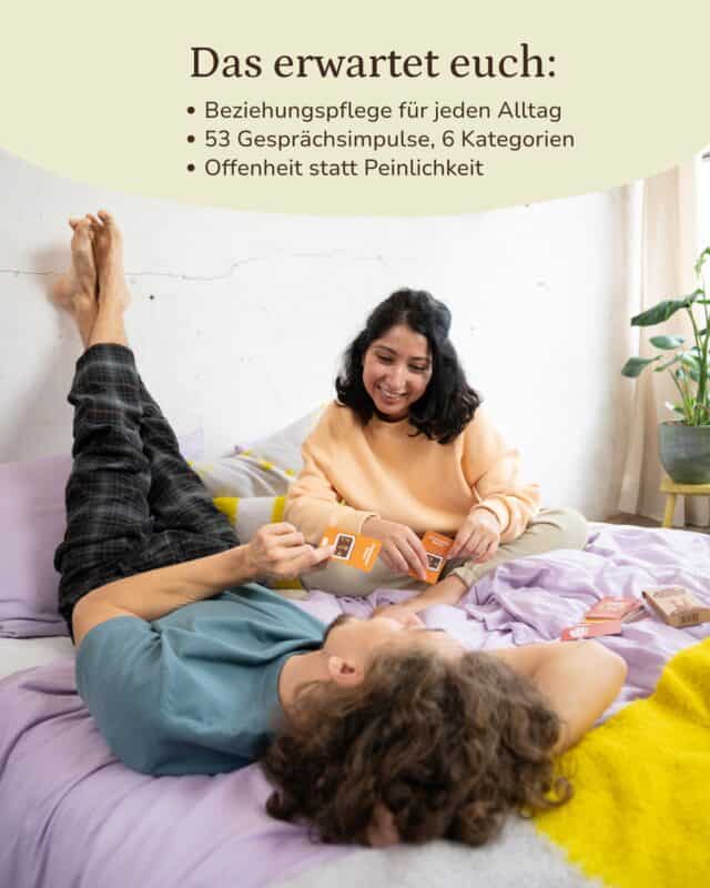 Ein Paar auf einem Bett mit lila und gelber Bettwäsche: Sie sitzt lachend im apricotfarbenen Sweater und hält zwei Fragekarten, er liegt quer auf dem Rücken, die Beine an der Wand; daneben Schachtel und Karten. Eingeblendet: „Das erwartet euch: Beziehungspflege für jeden Alltag · 53 Gesprächsimpulse, 6 Kategorien · Offenheit statt Peinlichkeit“