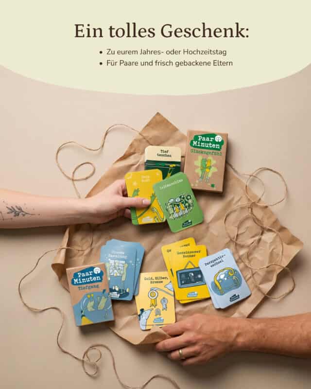 Auf Kraftpapier liegen die Schachteln PaarMinuten Glücksgefühl und PaarMinuten Tiefgang mit aufgefächerten Karten in Gelb, Grün und Blau; zwei Hände greifen von links und rechts ins Bild