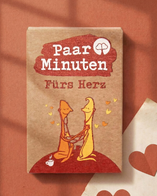 Schachtel PaarMinuten Fürs Herz auf terrakottafarbenem Untergrund, daneben Papier mit roten Herzen