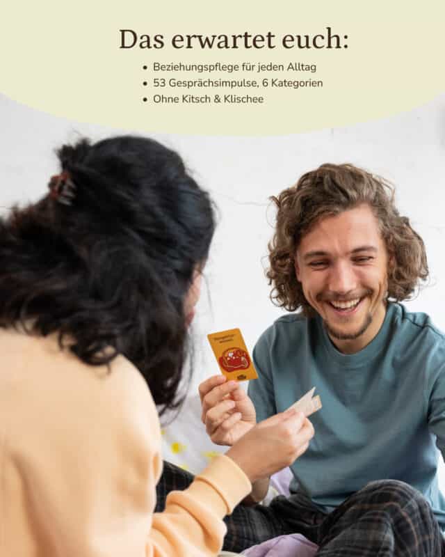 Ein Mann sitzt lachend auf einem Bett und hält eine gelbe Karte der Kategorie „Perspektivwechsel“ hoch, ihm gegenüber eine Frau von hinten mit orangefarbenem Pullover