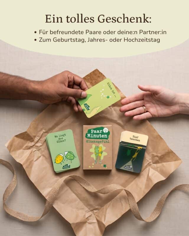 Von oben: zwei Hände reichen einander eine hellgrüne Karte „Glück ist …“ über Kraftpapier; darunter liegen die Schachtel PaarMinuten Glücksgefühl und zwei Kartenstapel der Kategorien „Wo liegt das Glück?“ und „Tief tauchen“, daneben ein Geschenkband. Eingeblendet: „Ein tolles Geschenk: Für befreundete Paare oder deine:n Partner:in · Zum Geburtstag, Jahres- oder Hochzeitstag“