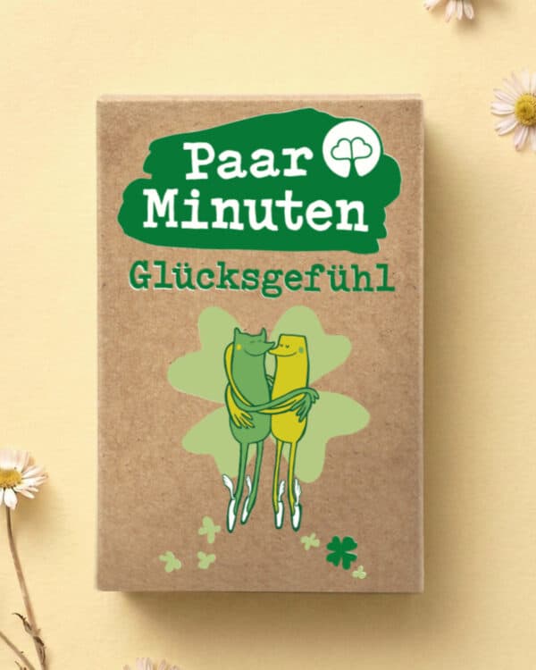 Schachtel PaarMinuten Glücksgefühl auf gelbem Untergrund mit Gänseblümchen, darauf zwei sich umarmende Figuren vor einem Kleeblatt