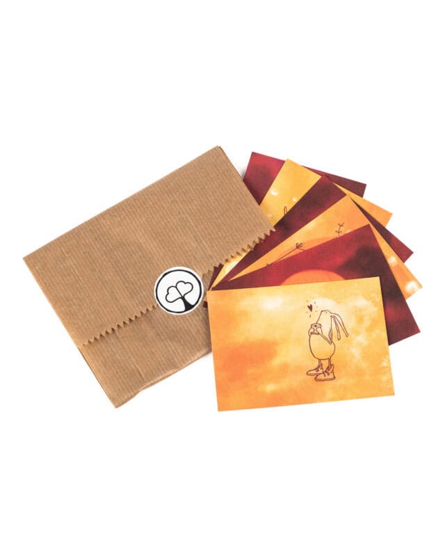 Vor weißem Hintergrund liegt eine flache braune Kraftpapiertüte mit gezacktem Rand, verschlossen mit einem runden weißen Aufkleber mit dem Paarzeit-Logo, einem Baum mit zwei Herzen als Krone. Rechts daneben sind sechs kleine Postkarten fächerförmig ausgelegt, alle in warmen Aquarelltönen von Gelb über Orange bis Dunkelrot; die vorderste Karte zeigt eine gezeichnete Figur mit langen Ohren und Turnschuhen, die etwas in den Händen hält, darüber ein kleines Herz.