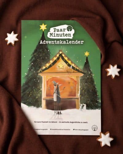 Der grüne PaarMinuten Adventskalender 2026 liegt auf braunem Strickstoff zwischen Zimtsternen; die Illustration zeigt ein Paar vor einer beleuchteten Weihnachtsmarktbude zwischen verschneiten Tannen, darunter „Für eure Paarzeit im Advent – 24 wertvolle Augenblicke zu zweit“.