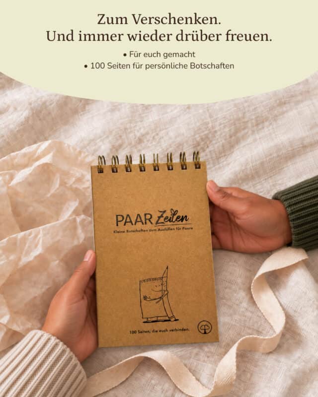 Zwei Hände halten den PaarZeilen-Block mit Kraftpapier-Umschlag über einer hellen Leinendecke, daneben liegen zerknittertes Seidenpapier und ein cremefarbenes Band. Die Person trägt einen hellen Strickpullover, von rechts reicht ein dunkelgrüner Strickärmel ins Bild. Auf dem Umschlag stehen „PAAR Zeilen“, „Kleine Botschaften zum Ausfüllen für Paare“ und „100 Seiten, die euch verbinden.“ mit der Zeichnung eines Notizblocks, der einen Bleistift umarmt. Eingeblendet oben in einer hellen Welle: „Zum Verschenken. Und immer wieder drüber freuen.“ · „Für euch gemacht“ · „100 Seiten für persönliche Botschaften“.