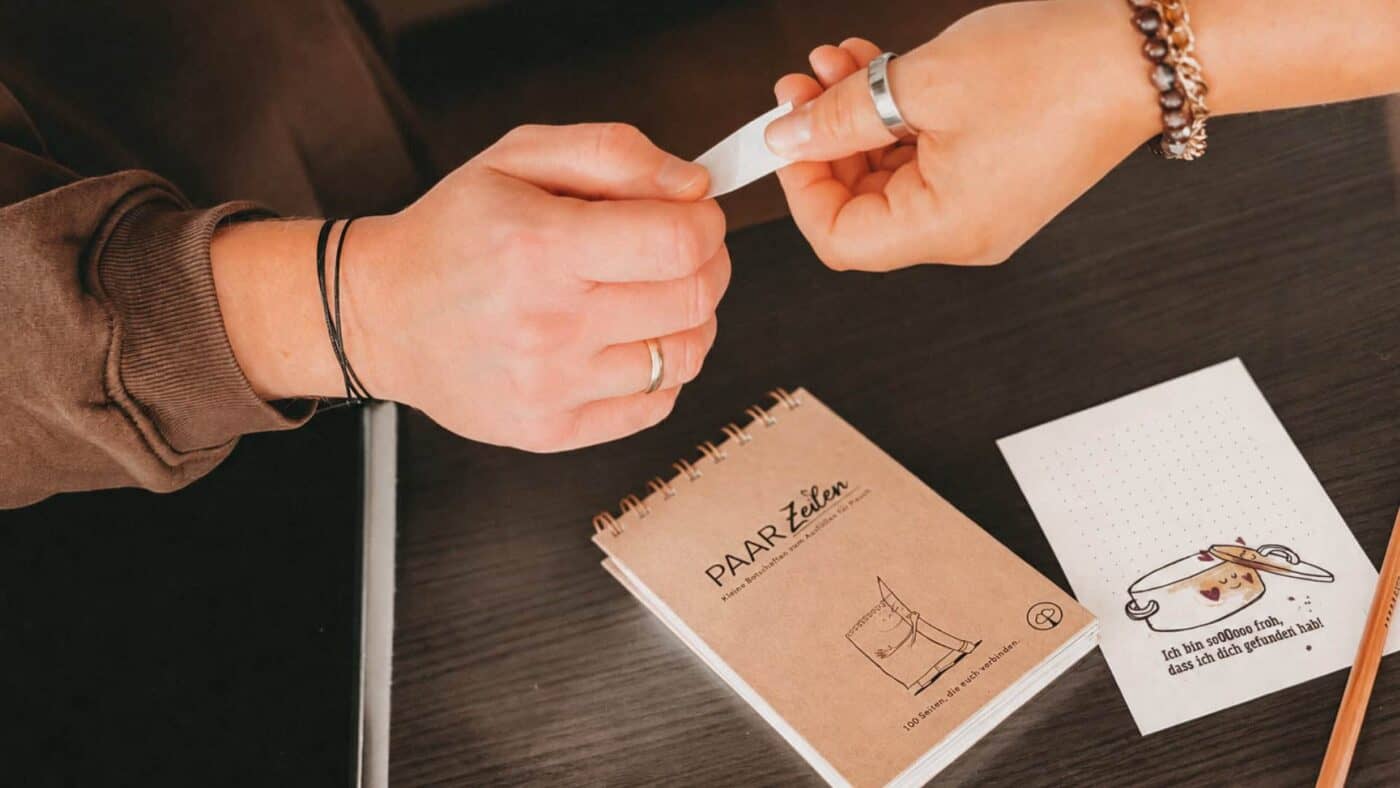 Zwei Hände reichen sich über einem dunklen Holztisch einen abgetrennten Papierstreifen weiter. Darunter liegt der PaarZeilen-Block mit Kraftpapier-Umschlag und Spiralbindung, daneben eine ausgefüllte Seite mit einem gezeichneten Topf, dessen Deckel lächelnd auf ihm sitzt, und ein Bleistift. Auf dem Umschlag: „PAAR Zeilen · Kleine Botschaften zum Ausfüllen für Paare · 100 Seiten, die euch verbinden.“ Auf der Seite: „Ich bin sooooo froh, dass ich dich gefunden hab!“