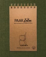 Der PaarZeilen-Block liegt hochkant und frontal fotografiert auf einem olivgrünen Leinenstoff, oben die goldene Spiralbindung. Der Umschlag ist aus Kraftpapier und schwarz bedruckt mit dem Schriftzug „PAAR Zeilen“ und dem Untertitel „Kleine Botschaften zum Ausfüllen für Paare“. Darunter ist ein gezeichneter Notizblock mit Gesicht zu sehen, der einen lächelnden Bleistift umarmt. Unten steht „100 Seiten, die euch verbinden.“, rechts daneben das Paarzeit-Baumzeichen im Kreis.