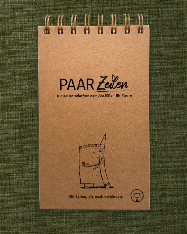 Der PaarZeilen-Block liegt hochkant und frontal fotografiert auf einem olivgrünen Leinenstoff, oben die goldene Spiralbindung. Der Umschlag ist aus Kraftpapier und schwarz bedruckt mit dem Schriftzug „PAAR Zeilen“ und dem Untertitel „Kleine Botschaften zum Ausfüllen für Paare“. Darunter ist ein gezeichneter Notizblock mit Gesicht zu sehen, der einen lächelnden Bleistift umarmt. Unten steht „100 Seiten, die euch verbinden.“, rechts daneben das Paarzeit-Baumzeichen im Kreis.