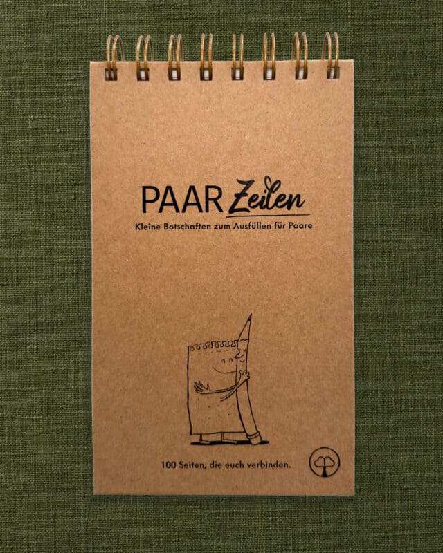Der PaarZeilen-Block liegt hochkant und frontal fotografiert auf einem olivgrünen Leinenstoff, oben die goldene Spiralbindung. Der Umschlag ist aus Kraftpapier und schwarz bedruckt mit dem Schriftzug „PAAR Zeilen“ und dem Untertitel „Kleine Botschaften zum Ausfüllen für Paare“. Darunter ist ein gezeichneter Notizblock mit Gesicht zu sehen, der einen lächelnden Bleistift umarmt. Unten steht „100 Seiten, die euch verbinden.“, rechts daneben das Paarzeit-Baumzeichen im Kreis.