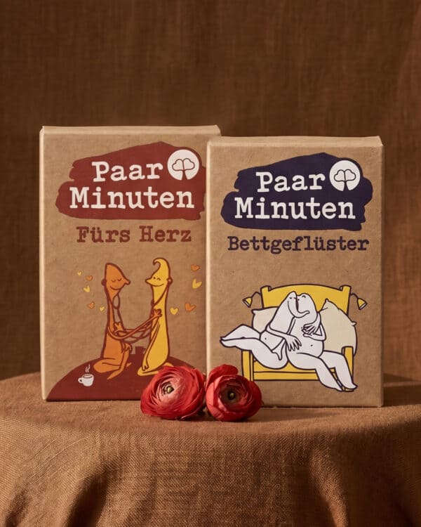 Die beiden Kraftpapier-Schachteln PaarMinuten Fürs Herz, mit einem illustrierten Paar, das sich an den Händen hält, und PaarMinuten Bettgeflüster, mit einem illustrierten Paar in einem gelben Bett, stehen auf einem runden Tisch mit braunem Leinentuch; davor liegen zwei rote Ranunkeln.