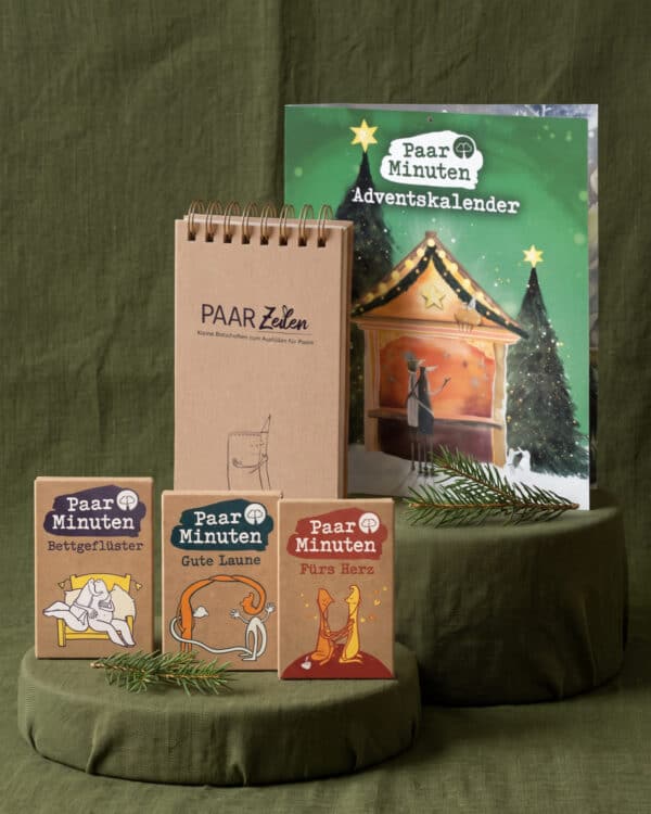 Fünf Paarzeit-Produkte auf zwei runden, mit grünem Stoff bezogenen Podesten vor olivgrünem Tuch: hinten der grüne PaarMinuten Adventskalender 2026 und der PaarZeilen-Block aus Kraftpapier mit Spiralbindung, vorn die Schachteln PaarMinuten Bettgeflüster, Gute Laune und Fürs Herz; dazwischen Tannenzweige.