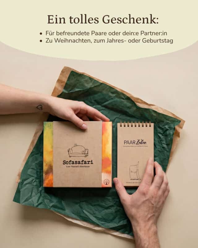 Zwei Hände legen die Sofasafari-Box und den PaarZeilen-Block auf grünes Seidenpapier, das auf zerknittertem Packpapier vor hellem Grund liegt; am Handgelenk der oberen Hand ein kleines Herz-Tattoo. Eingeblendet: „Ein tolles Geschenk: Für befreundete Paare oder dein:e Partner:in. Zu Weihnachten, zum Jahres- oder Geburtstag.“
