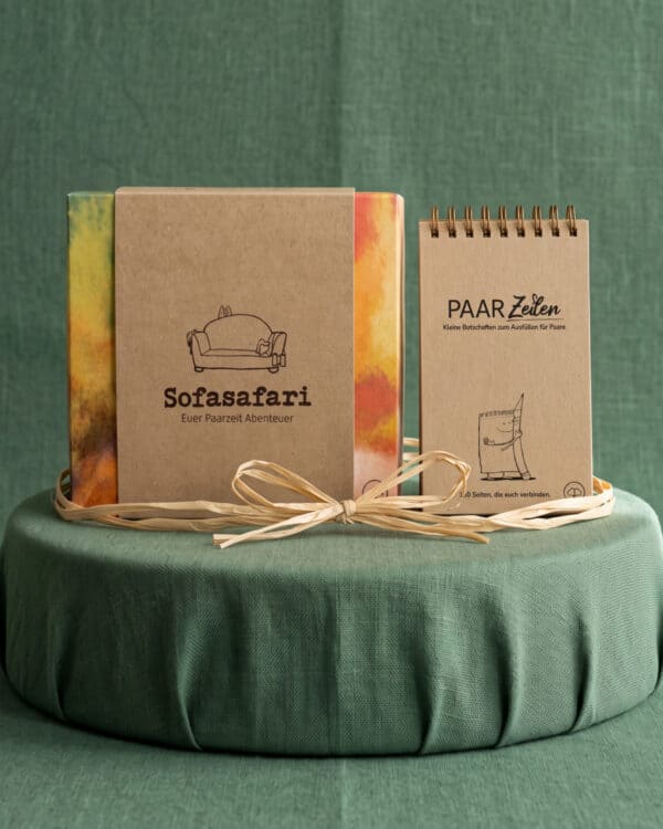 Die Sofasafari-Box mit Kraftpapier-Banderole und bunt gefärbten Seiten steht neben dem PaarZeilen-Block mit Spiralbindung auf einem runden, mit grünem Stoff bezogenen Podest vor grünem Tuch; um beide ist eine Bastschleife gebunden.